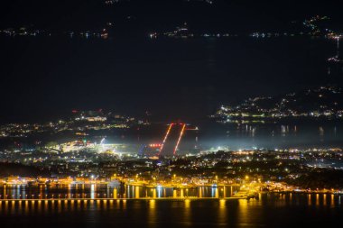 Gece aydınlık bir kıyı şehrini görüntüleyen bir hava görüntüsü. Liman ışıkları suya yansıyor. Parlak ve canlı bir manzara yaratıyor..