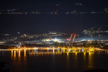 Bir gece manzarası, suların ötesinde manzarayı aydınlatan ışıklarla bir şehir sergiliyor. Karanlık su, canlı şehir manzarasını yansıtıyor..