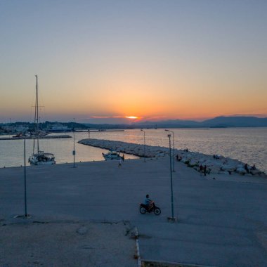 Sahil manzaralı sakin bir manzarada, uzaklardaki dağ manzaralı günbatımında motorsikletliler gibi rıhtımın yakınına demirlemiş tekneler sergileniyor..