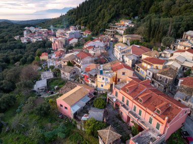 Manzaralı bir hava manzarası, yemyeşil bir yamaçta yuva yapmış bir köyü yakalıyor, kırmızı kiremit çatılı büyüleyici evleri sergiliyor, hepsi bulutlu bir gökyüzünün altında..