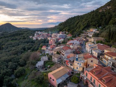 Yüksek açılı bir çekim renkli evleri olan, Alacakaranlık 'ta uzak bir denizi gören yeşil bir yamaçta birleşen antika bir köy sunar..