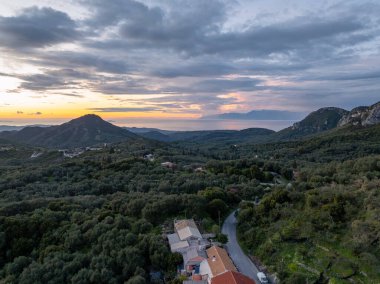 Bulutlu bir gökyüzünün altında denize uzanan ağaçlarla kaplı dağlık bir manzara. Yeşillikler arasında evler ve bir yol yuvası.