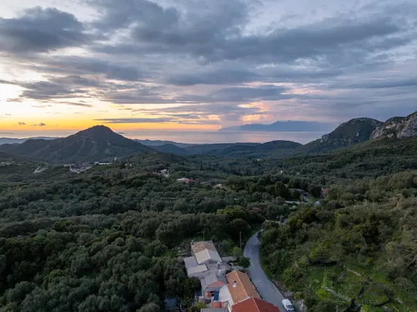 Bulutlu bir gökyüzünün altında denize uzanan ağaçlarla kaplı dağlık bir manzara. Yeşillikler arasında evler ve bir yol yuvası.