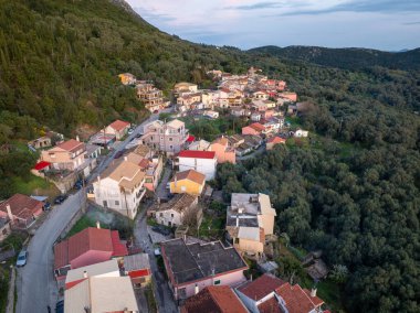 Yüksek açılı bir manzara, yemyeşil ağaçlarla kaplı tepelerde, kırmızı ve beyaz çatıları olan çeşitli binaları gösteren bir Avrupa köyünü gösteriyor..