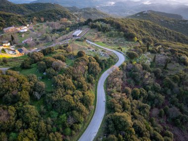 Canlı, yeşil bir yamaçtan geçen ve binaları doğal manzaranın güzelliğine bağlayan bir yolun havadan görünüşü..