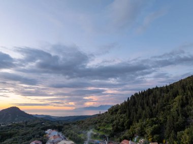 Manzaralı bir manzarada ağaçların arasına yerleşmiş bir yamaç köyü, sakin bir okyanus ve alacakaranlıkta bulutlu bir gökyüzünün altında görünen dağlar var..