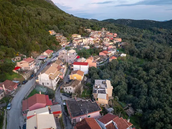 Yüksek açılı bir manzara, yemyeşil ağaçlarla kaplı tepelerde, kırmızı ve beyaz çatıları olan çeşitli binaları gösteren bir Avrupa köyünü gösteriyor..