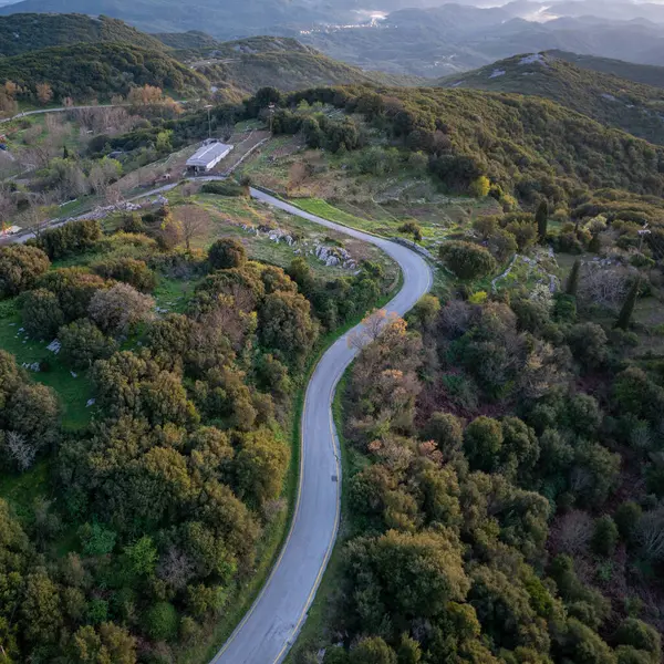 Yüksek açılı bir manzara yeşil tepeler boyunca kıvrımlı bir yol gösteriyor, ağaçlarla süslenmiş, ve arka planda sisli dağlar.. 