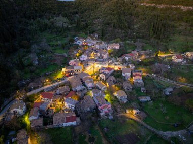 Manzaralı bir hava görüntüsü vadide yuva yapmış büyüleyici bir köyü yakalıyor. Binalar, alacakaranlıkta yeşil tepelere karşı sıcak bir şekilde parlıyor..