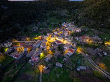 Alacakaranlıkta ağaçların arasına kurulmuş küçük bir köyü hava manzarası yakalıyor. Aydınlatılmış sokak lambaları ve evler yeşil ortamda rahat bir atmosfer yaratıyor..