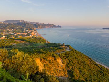 Kıyı kasabasına bakan yeşil bir yamaçtan geniş bir manzara ve günbatımında turkuaz okyanus, arka planda yuvarlanan tepeler.