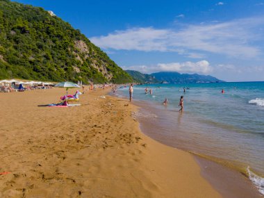 Güneşli bir günde yakalanan turkuaz suyla dolu geniş kumlu bir sahil yemyeşil bir tepenin yanındaki odak noktadır. İnsanlar rahatlıyor ve yüzüyor..