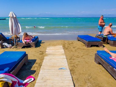 Canlı bir plaj sahnesi. İnsanlar suyun kenarındaki sandalyelerde uzanıyorlar. Açık gökyüzü bir yaz gününde turkuaz denize bakar..