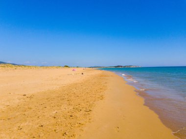 Bu çekim, açık mavi gökyüzünün altında turkuaz deniz bulunan kumlu bir sahili gösteriyor. Sahne sakin bir günde huzur ve rahatlık yayar..