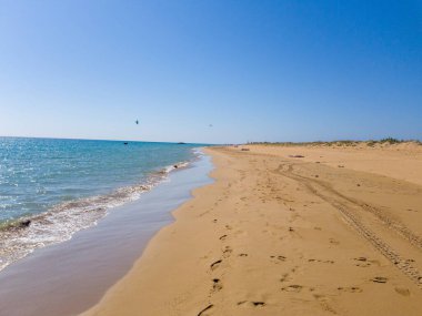 Parlak, bulutsuz bir gökyüzünün altında el değmemiş kumlu bir sahil berrak mavi denizle buluşur. Lastik izleri kumu işaret ediyor, uzak kumullara doğru gidiyor..