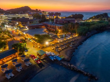 Alacakaranlıktaki canlı bir sahil kasabasının havadan görünüşü. Ilık ışıklar binaları, plaj sandalyelerini ve pedallı tekneleri aydınlatır, rahat bir ortam yaratır..