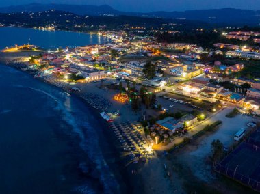 Geceleri, parlak ışıklı binalar, plaj şemsiyeleri, karanlık okyanus dalgaları ve uzak dağların yer aldığı bir sahil kasabasının havadan görünüşü..