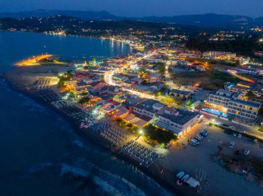 Gece hava manzarası aydınlık binalar, kumlu bir sahil ve karanlık bir gökyüzünün altında sakin bir deniz olan bir kıyı kasabasını gösteriyor. Arkaplanda dağlar.