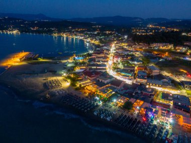 Geceleri deniz manzarası, aydınlık binalar, şemsiyeli bir plaj ve karanlık bir gökyüzünün altında sakin bir okyanus..