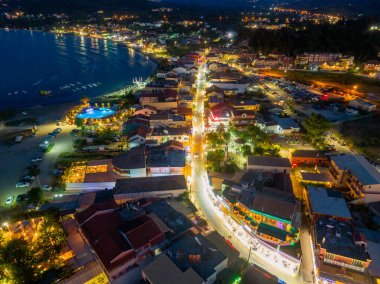 Hava gece manzarasında, aydınlık binalar, sokak lambaları, kumsal ve alacakaranlıkta su özellikleriyle parıldayan bir kıyı kasabası görülüyor..