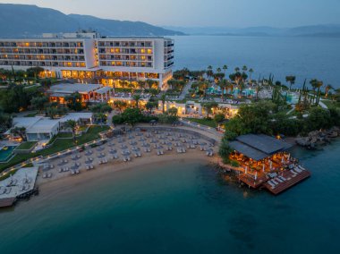 Büyük bir binası, şemsiyeli plajı ve sıcacık ışıklarla aydınlatılmış iskele restoranı olan lüks bir tatil köyünün havadan alacakaranlık manzarası..