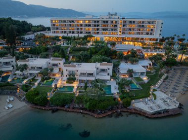 Havadan çekilen görüntüler alacakaranlıktaki lüks bir tatil köyünü gösteriyor. Parlayan binalar, havuzlar ve sakin deniz boyunca şezlongları olan bir plaj..