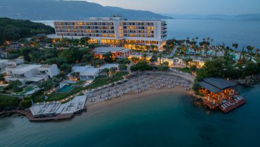 Şemsiyelerle kaplı kumlu bir sahil, büyük bir otel ve yemyeşil bir çevreye sahip, gece karanlığında lüks bir tatil köyünün havadan görünüşü..