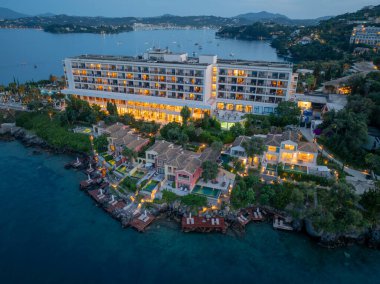 Lüks bir sahil beldesinin havadan çekilmiş fotoğrafı. Bir otel ve birden fazla villa var. Denize bakıyor ve birçok ışık yanıyor. Alacakaranlık ışıkları.