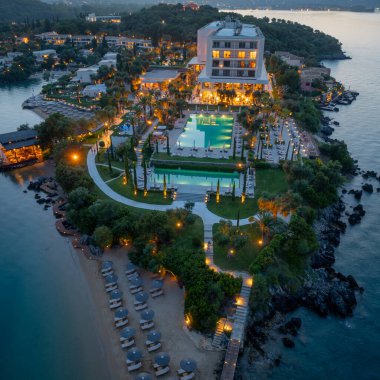 Deniz kıyısındaki bir tatil köyünün sergilendiği panoramik bir akşam sahnesi, aydınlatılmış havuzlar, kumlu bir plaj ve su kenarındaki lüks konaklama yerleri.
