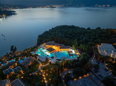 Gökyüzü alacakaranlık altında, büyük bir su kütlesinin yanında, ışıklarla aydınlatılmış, ağaçlarla kaplı bir sahilde bir tatil köyü yakalıyor..