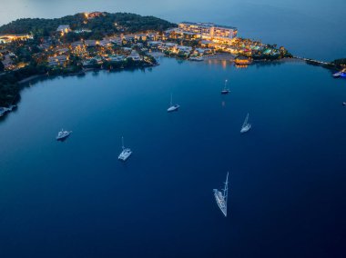 Alacakaranlık saatlerinde kıyı şeridindeki parlak ışıklandırılmış bir tesis tarafından çerçevelenen sakin bir körfezde demirleyen tekneleri bir hava manzarası yakalıyor..
