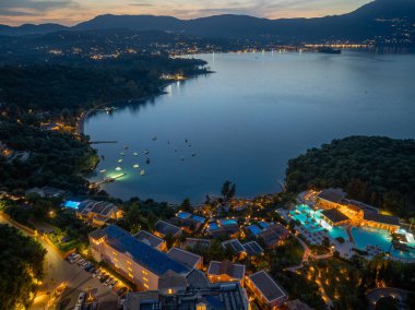 Bu hava görüntüsü, sahil şeridinde bir tatil beldesini çekiyor. Hafif ışıklar, binaları, havuzları ve dağ sırtlarının altındaki sakin körfezde bulunan tekneleri aydınlatıyor..