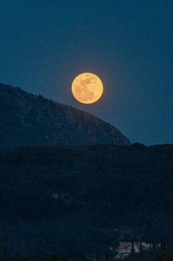 Dolunay gölgeli bir dağ sırtının ötesine yükselir, bulutlu bir akşamda aşağıdaki karanlık ormanda hafif bir parıltı bırakır..