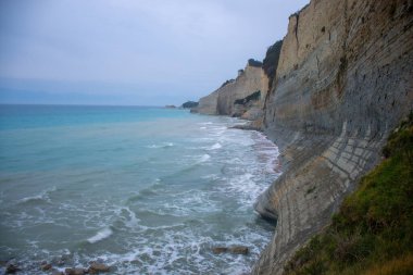 Uzun, katmanlı uçurumların bulutlu bir gökyüzü altında küçük dalgalarla turkuaz suya düştüğü dramatik kıyı manzarası; kayalık kıyı şeridi ve yeşil büyüme.