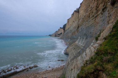 Sakin bir turkuaz deniz boyunca engebeli kıyı uçurumlarının yer aldığı, bulutlu bir gökyüzünün altında sakin, doğal bir manzara yaratan pitoresk bir manzara..
