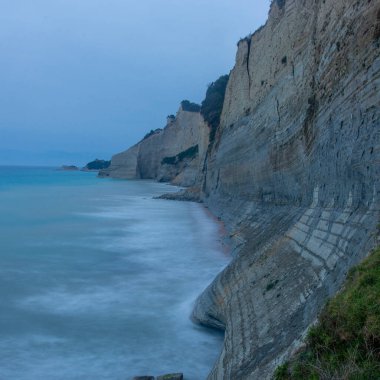 Uzun bir pozlama, katmanlı sahil kayalıklarının ve sakin denizin alacakaranlık gökyüzünün altında sakin bir manzarayı yakalar..