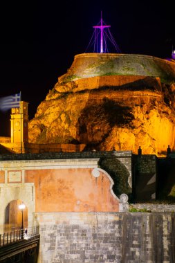 Geceleri aydınlatılmış yüksek bir kale, altında eski taş duvarlar, köprü ve üzerinde bayrak olan bir saat kulesi..