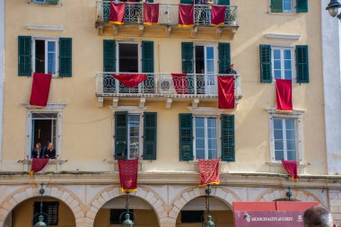 Açık sarı renkli bir bina, pencerelerden ve balkonlardan sarkan kırmızı bezlerle süslenir. Bu da özel bir olay ya da kutlama anlamına gelir..