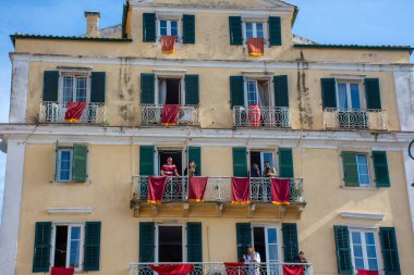 Sarı cepheli, yeşil panjurlu ve işlenmiş demir balkonlu çok katlı bir bina. İnsanlar kırmızı kumaşla balkonda..