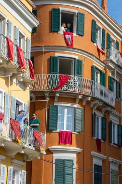 Bir şehir sahnesinde, kırmızı örtülerle süslenmiş, balkonlardan ve mermer binaların pencerelerinden canlı ve canlı bir atmosfer yaratan insanlar görülüyor..