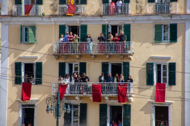 Bronzlaşmış dış duvarları, yeşil panjurları ve birçok balkonu olan çok katlı bir bina. Kırmızı perdeler binayı süslüyor..