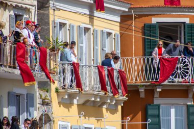 İnsanlar balkonlarda toplanır, bazıları geleneksel kıyafetler giyer, parlak renkli bir Avrupa şehrinde gün ışığı altında bir sokak kutlamasına tanıklık etmek için..