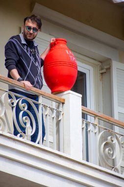 Dekoratif demirle beyaz boyanmış bir balkonda elinde büyük bir kırmızı kilden sürahi ve açık tenli güneş gözlüğü tutan bir adam var..
