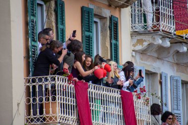 Kalabalık balkonda kutlama yapıyor, kırmızı kapları fırlatırken fotoğraf çekiyorlar. Binanın mavi panjurları ve kırmızı dekoratif afişi var..