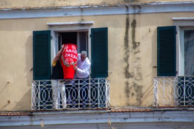İki adam büyük, kırmızı, dekore edilmiş bir vazoyu dikkatlice yeşil panjurlu bir balkona itti..