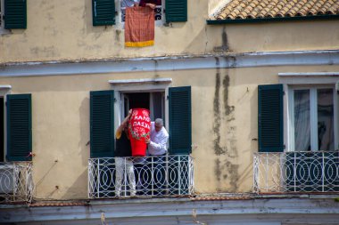 İnsanlar Paskalyayı balkondan kutluyor, büyük kırmızı bir vazo fırlatmaya hazırlanıyorlar. Yunanistan 'ın Korfu kentindeki yeşil panjurlu bina cephesi.