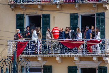 Bir grup balkonda duruyor, hareketli bir etkinliğin tadını çıkarıyorlar. Binanın yeşil panjurları ve iki kişinin taşıdığı büyük kırmızı bir vazosu var.. 
