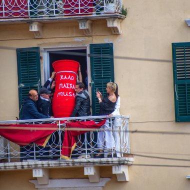 Birkaç kişi bir balkonda duruyor. Bir kadın gösterinin fotoğrafını çekerken onlar büyük, kırmızı bir vazoyu kapıdan geçiriyorlar..