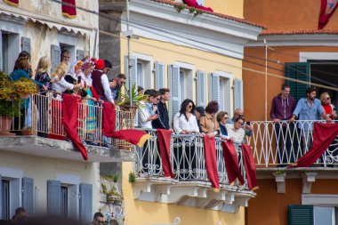 Bir grup insan, kırmızı pankartlarla süslenmiş renkli binaların balkonlarında canlı bir açık hava ortamında eğleniyor..