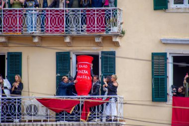 İnsanlar pencereden kırmızı bir tencere fırlatıyor. Balkonlarda insanlar var. Esrar Yunanistan 'da Paskalya için atılıyor.